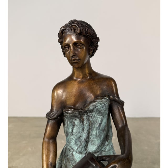 Refined figural bronze sculpture depicting a seated woman holding a vessel, rendered in a style inspired by classical...
