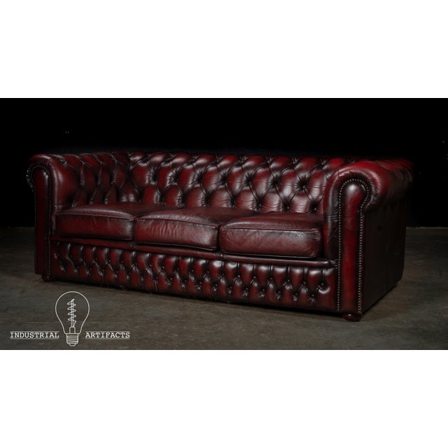 Classic Chesterfield sofa upholstered in richly colored oxblood leather with deep diamond tufting across the back, rolled...