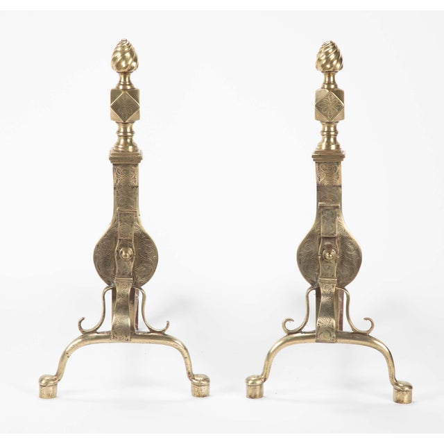 A fine pair of brass andirons with scrolling etched designs. The flame finials over diamond squares above a vase form body...