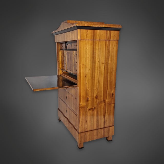 A Biedermeier-era secretary desk. The body is veneered in cherry wood with a continuous veneer pattern. It is topped by a...