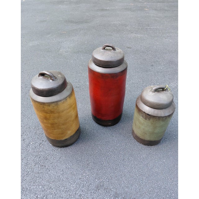 A charming set of three handcrafted stoneware lidded jars featuring earthy ochre, oxblood red, and soft sage green glazes...