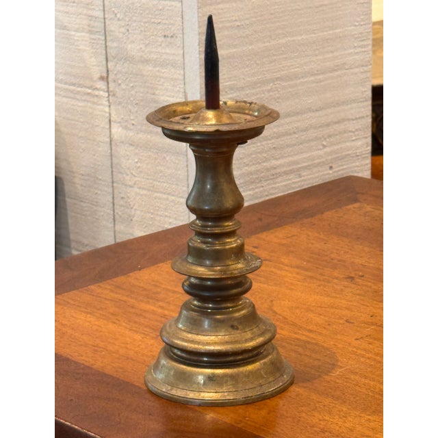 A single bronze pricket stick with a turned column and wide drip pan, raised on a stepped circular base. The surface shows...