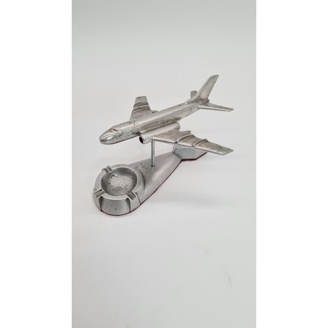 An original metal desk ashtray in the shape of a jet plane, mounted on a base with space for a cigarette. The item is...