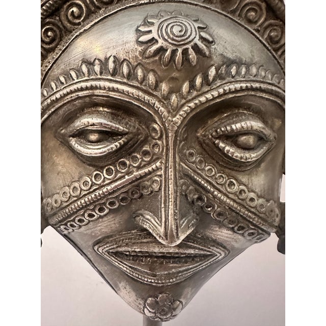 A unique metal sculpture featuring a stylized tribal face mounted on a conical base. The piece showcases intricate...