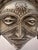 A unique metal sculpture featuring a stylized tribal face mounted on a conical base. The piece showcases intricate...