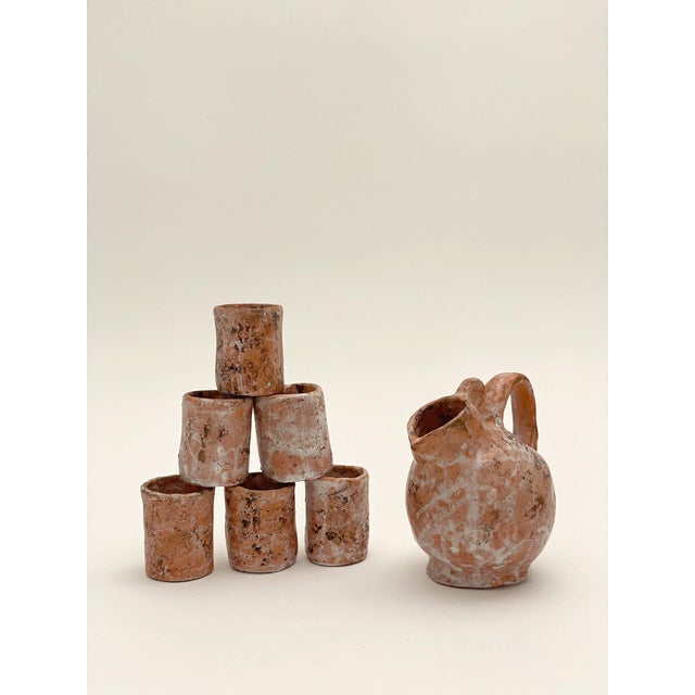A set comprising a jug and six small terracotta cups, finished with a white and pink engobe with a marbled effect. The...