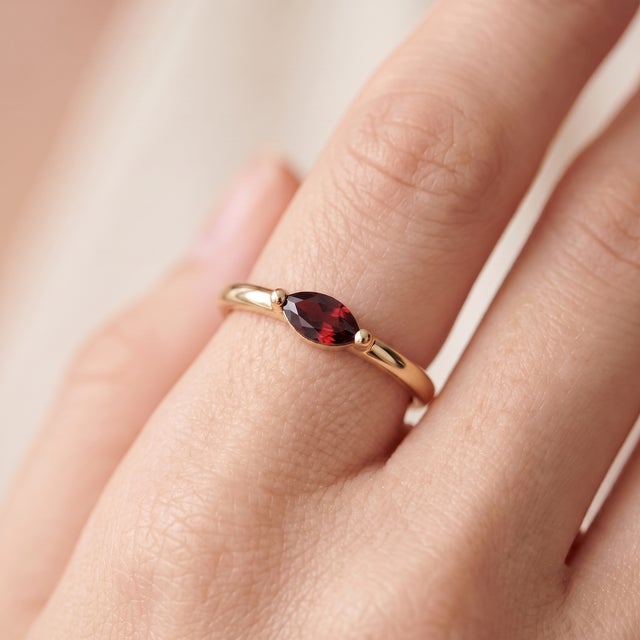 A dainty, modern solitaire featuring a rich red marquise-cut garnet set sideways in an east-west orientation, cradled by a...