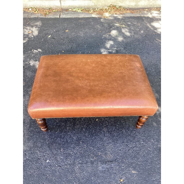 A handsome rectangular ottoman featuring warm cognac leather upholstery, turned wooden legs, and brass-tone nailhead trim....