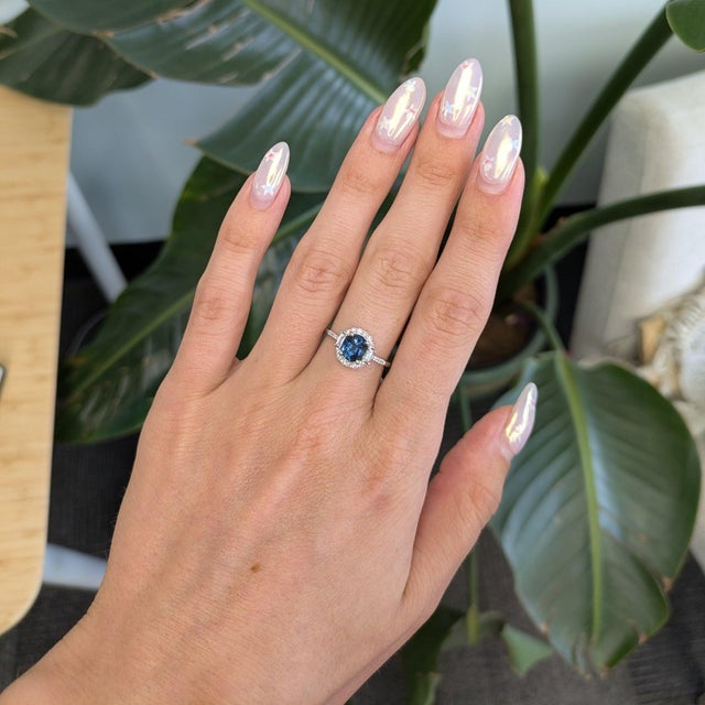 A captivating teal sapphire blends ocean blue and forest green at the center of this oval ring, framed by a halo of...