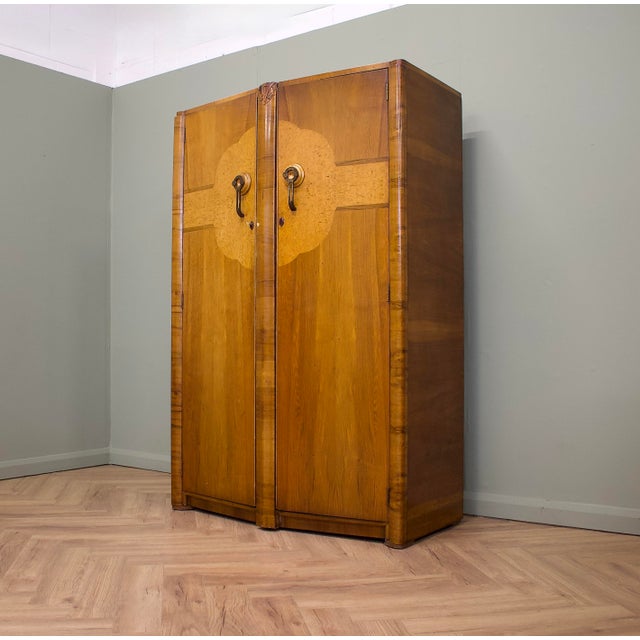 A striking 1930s Art Deco double wardrobe featuring figured walnut veneer, book-matched door panels, carved central...
