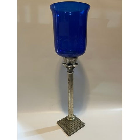 A single tall hurricane candle holder featuring a silver metal corinthian column base topped with a blue glass hurricane...
