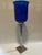 A single tall hurricane candle holder featuring a silver metal corinthian column base topped with a blue glass hurricane...
