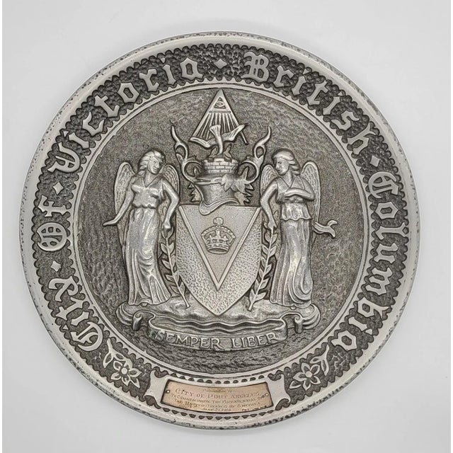 Here is a pewter plate depicting two angels flanking a crest, representing the city of Port Angeles, Washington, presented...