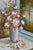 A lush and romantic floral still life depicting a generous bouquet of pink roses in a white vase set against a mullioned...