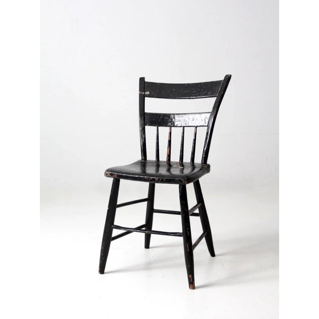 An antique primitive chair featuring a painted black plank seat, a half-spindle back, and splayed legs. Its understated...