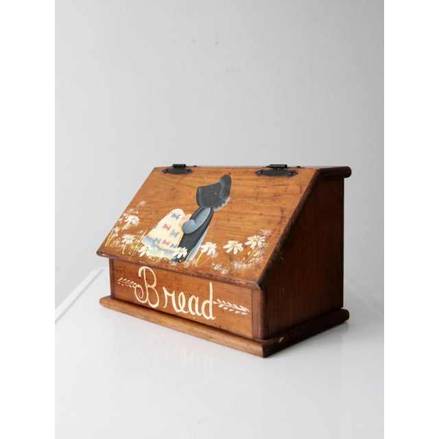 This vintage hand-painted wood bread box features a sloped lid, warm wood tone, and a Holly Hobbie style folk scene that...