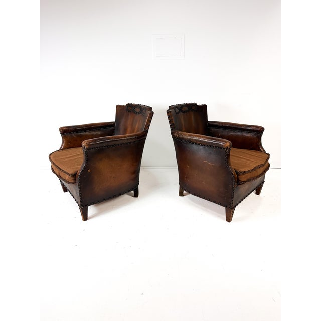 Pair of early 20th-century antique European club chairs. Original brown leather features a rich, time-earned patina with...