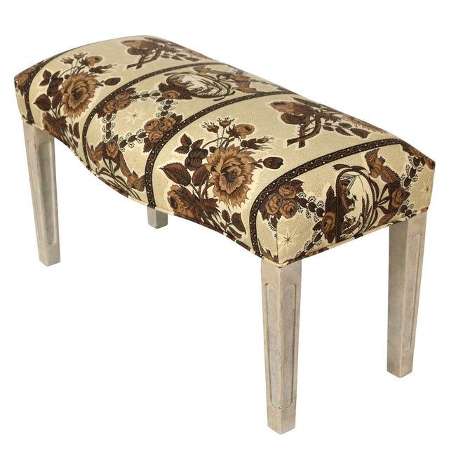 A charming upholstered bench with a curved seat covered in a warm-toned floral textile featuring a structured, repeating...