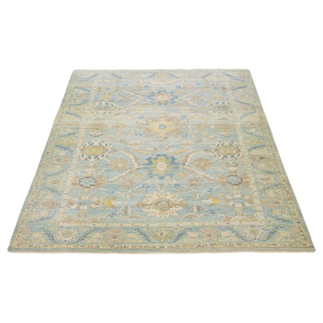 This hand-knotted wool rug has a light blue background adorned with yellow, beige, green, and brown all-over designs. This...