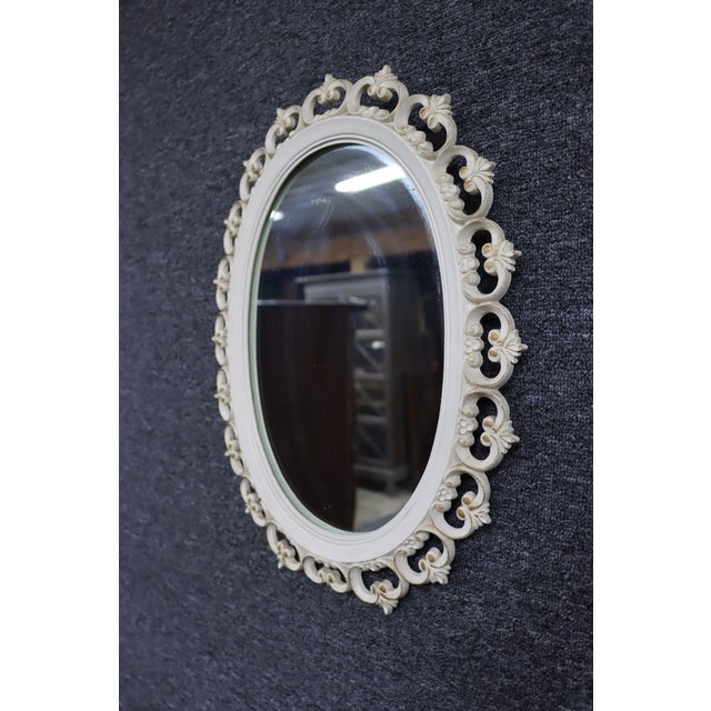 Oval wall mirror featuring an elegant pierced-cut frame with repeating scroll and fleur-de-lis motifs. Finished in white...