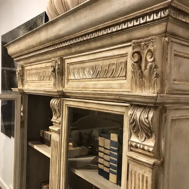 Wood carved vitrine featuring a light distressed finish with carved acanthus leaves and crown moulding, two lower drawers,...