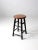 Vintage wooden stool featuring a round seat supported by turned legs finished in black. The contrast between the natural...