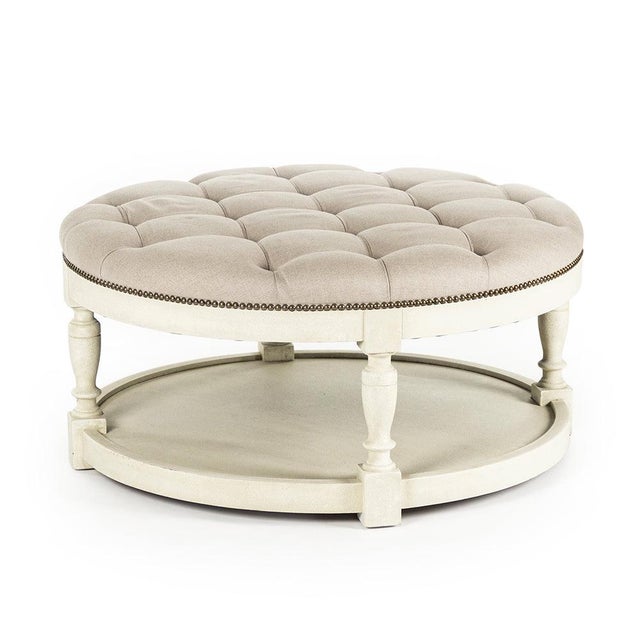 The Tammy Ottoman from Zentique is a round tufted ottoman in natural linen with brass nailhead trim on a distressed ivory...