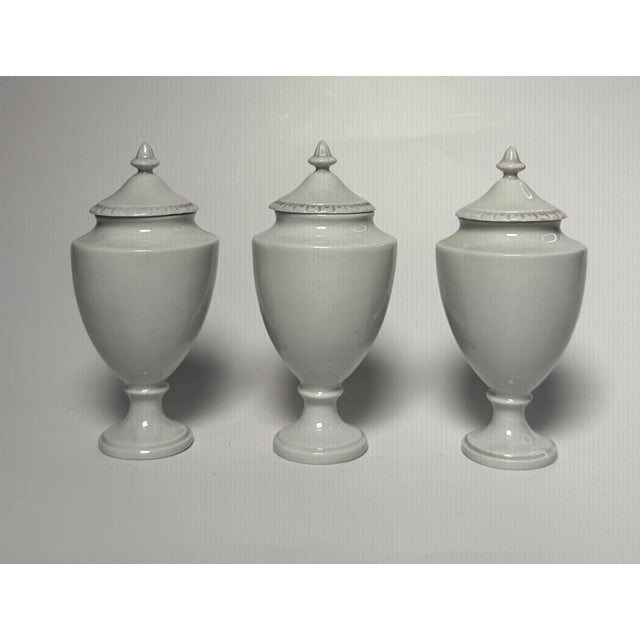 Clean, glossy white porcelain in a neoclassical style adorn this small set of porcelain urns. Each vessel has a lid with...