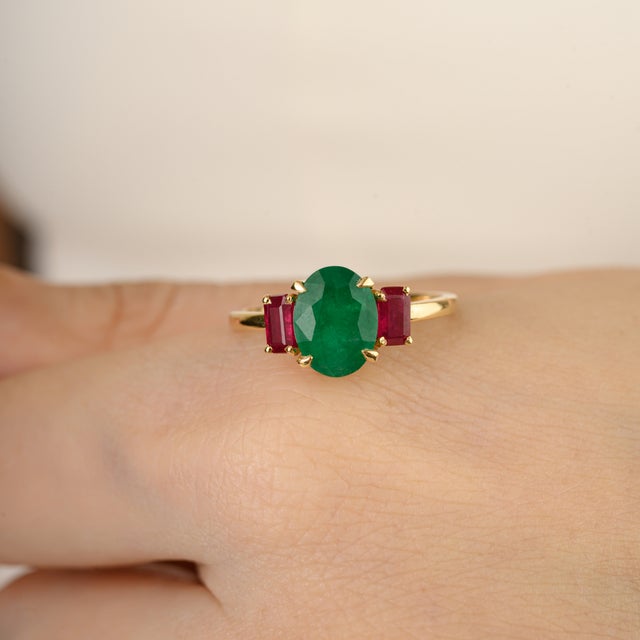 This elegant three-stone ring showcases an oval-cut 2.1Ct natural emerald at the center, flanked by two baguette-cut...