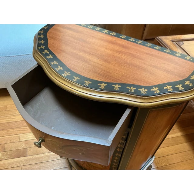 French-inspired demilune cabinet reproduction featuring ornate hand-painted gold fleur-de-lis and scrollwork motifs on a...