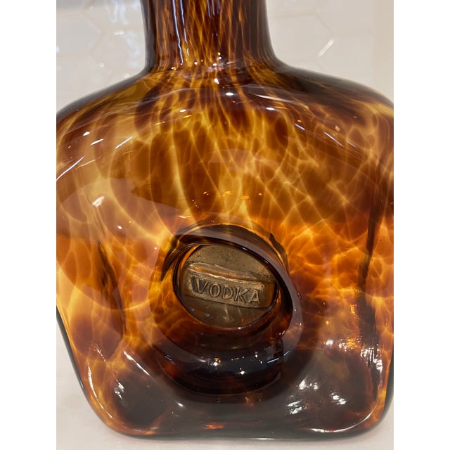 Hand blown faux tortoiseshell glass decanter with decorative brass stopper and brass inserts. The front insert has a Vodka...
