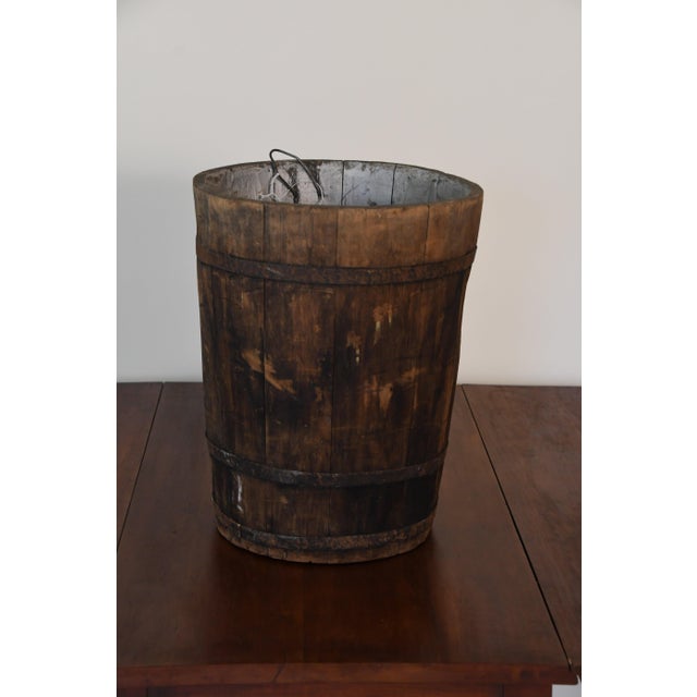 A wooden sap bucket of the kind once hung on maple trees each spring to collect sap. Built from staves held with metal...