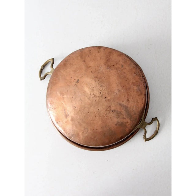 An antique heavy-gauge copper pan featuring twin brass handles and a gently rounded base. The substantial copper...