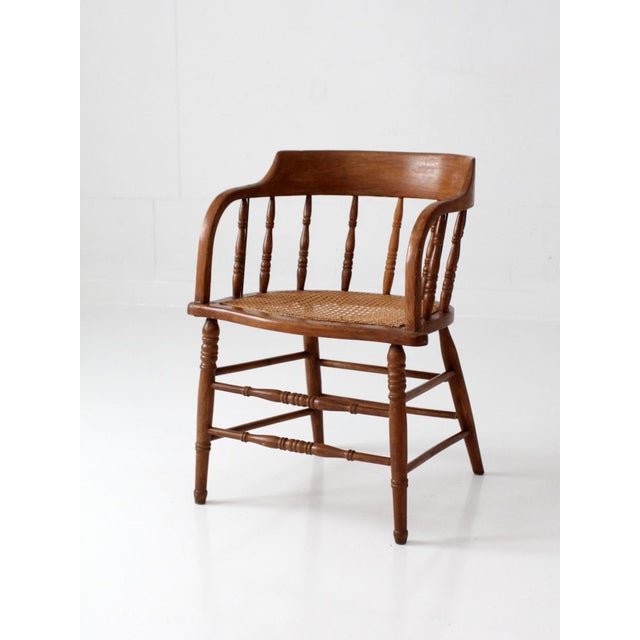 This antique cane seat captain’s chair features a curved crest rail with shaped arms, turned spindle supports, and a hand-...