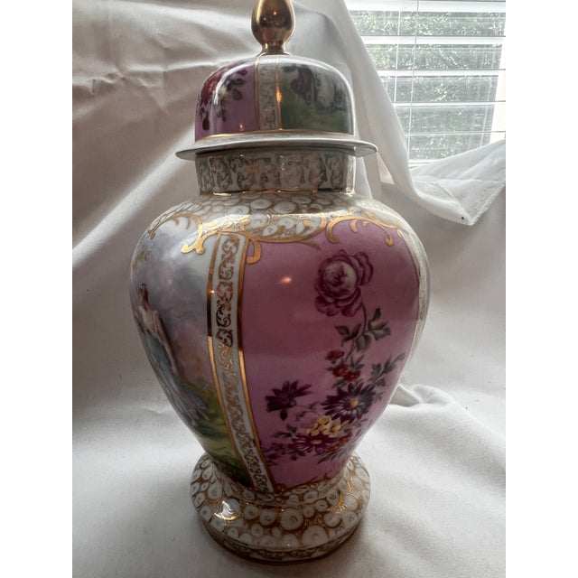 A striking pair of vintage porcelain covered urns featuring romantic pastoral scenes, floral panels, and ornate gilt...