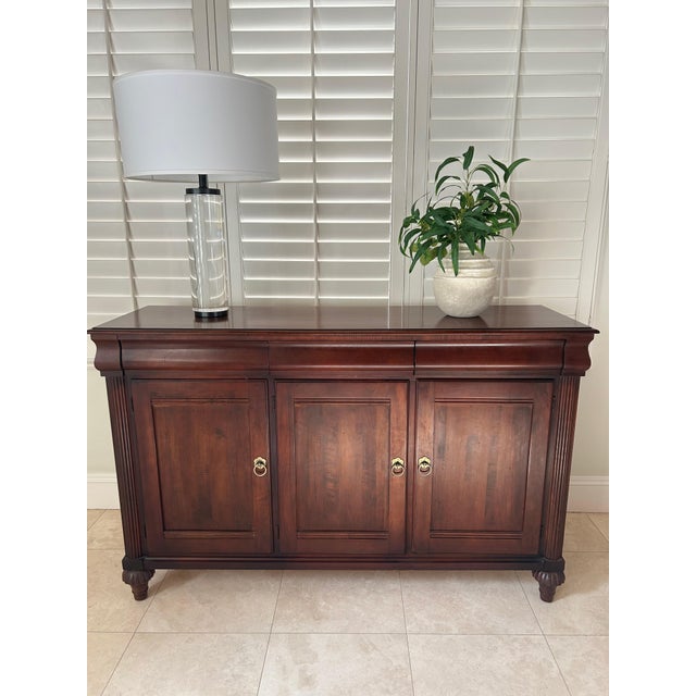 This classic British Colonial sideboard / buffet is made by Ethan Allen. Crafted from maple in warm Cinnabar stain with...