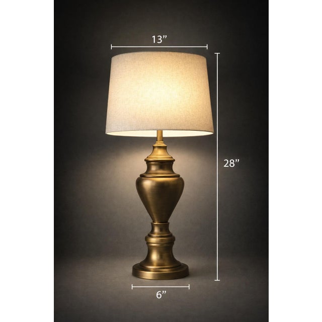 A classic antiqued brass urn table lamp with a large linen drum shade and elegant neoclassical silhouette. Warm brass...