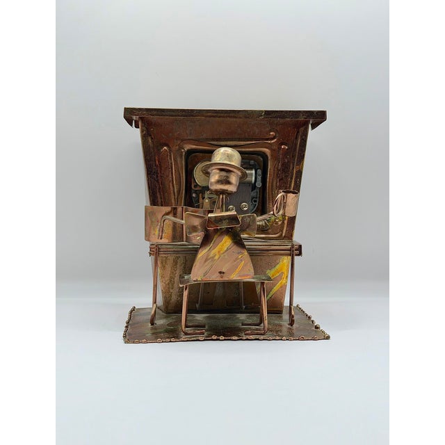 A vintage copper-toned music box featuring a stylized piano player seated at a grand piano, rendered in sculptural metal....