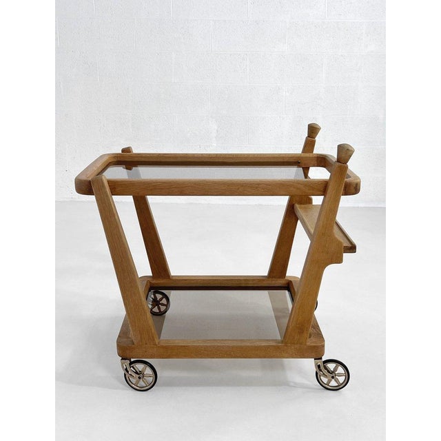 French design bar cart by Guillerme et Chambron, circa 1960, crafted from oak with smoked glass shelves. It features a...