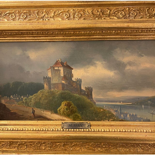 Decorative pair of framed landscape paintings by M. Day, each depicting a romantic European riverside castle scene. The...