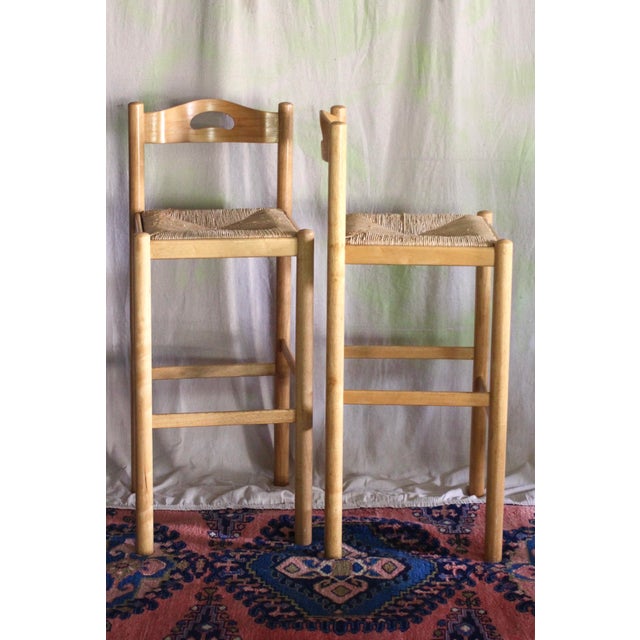 Set of 2 stools in the style of Hank Lowenstein. Blonde light wood in a great 70s style. Seats are woven rush. Cut out...