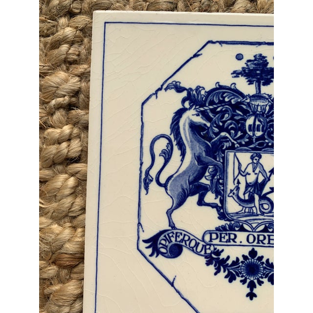 A classic Dutch Delft-style hand painted blue and white porcelain tile featuring an intricately rendered heraldic coat of...