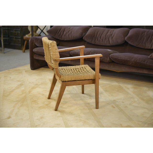 A wooden armchair with straight armrests, typical of post-war French furniture, designed for sturdiness and ease of...