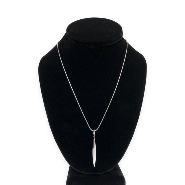 A sleek, elongated feather pendant in polished 18K white gold circa 1990's, suspended from a smooth round snake chain. The...
