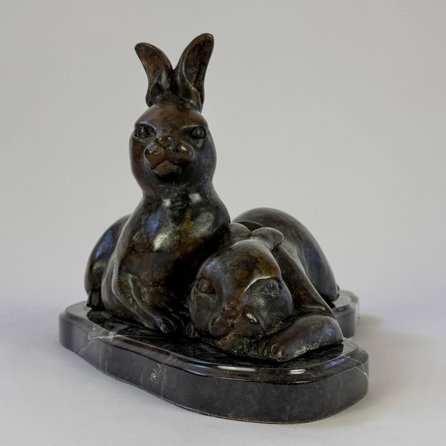 This bronze sculpture depicts a resting hare accompanied by a companion rabbit nestled beside it, creating a peaceful and...