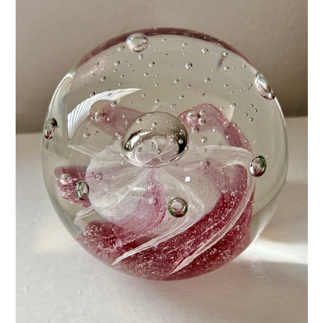 Beautiful round clear glass paperweight with a soft pink and white swirl inside. Small controlled bubbles float around a...