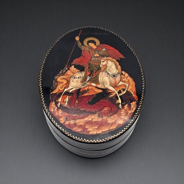 An exquisitely hand-painted Russian lacquer miniature box depicting Saint George astride a white horse, triumphing over...