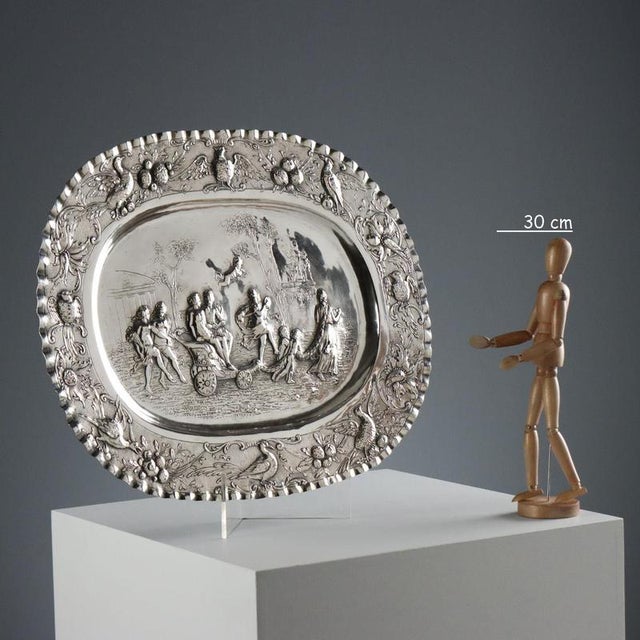 Embossed, chased, and burin-worked silver tray. At the center is a scene of a festive procession. The brim features a...