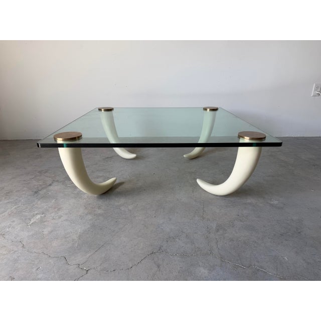 Striking vintage coffee table featuring four sculptural faux elephant tusk legs supporting a thick glass top with solid...