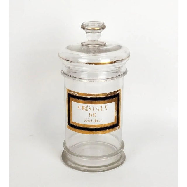 Pharmacy jar and its glass lid bearing the inscription "Cristaux de soda" in gold on a white background.